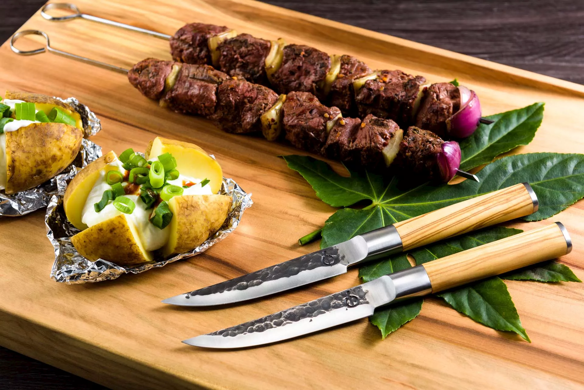 Inspirationsbild två Köttknivar forged. på skärbräda med grillspett och potatis. Handtag i olivträ.